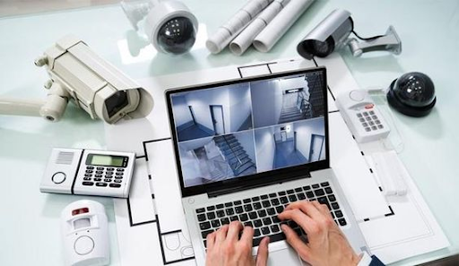 Person monitoring security camera footage on a laptop surrounded by surveillance equipment.